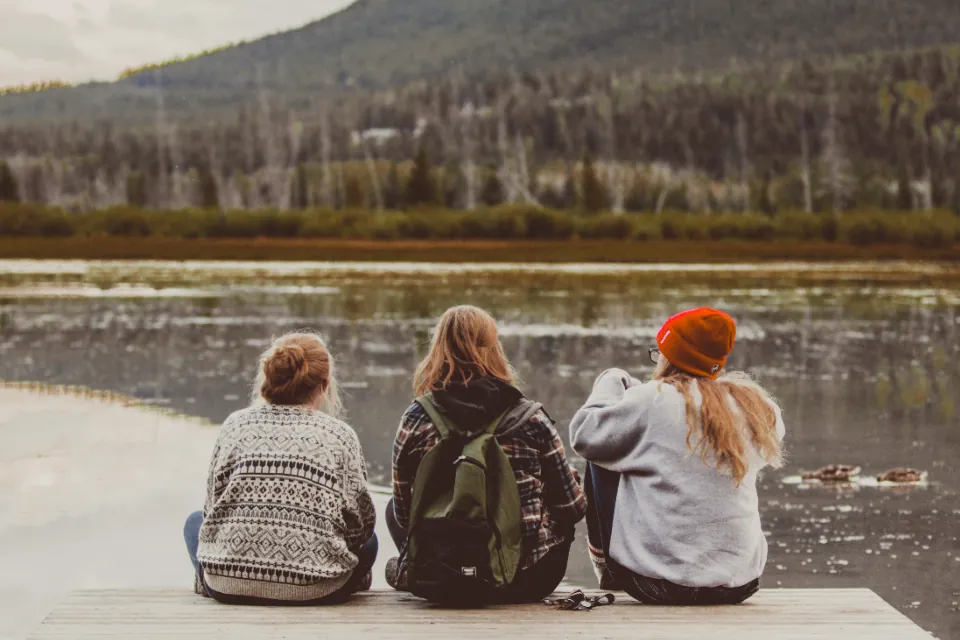 Três jovens sentados lado a lado à beira de um lago, de costas, conversando