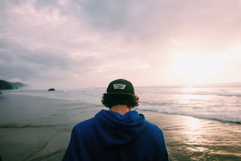 Jovem de boné, de costas, olhando o mar ao entardecer