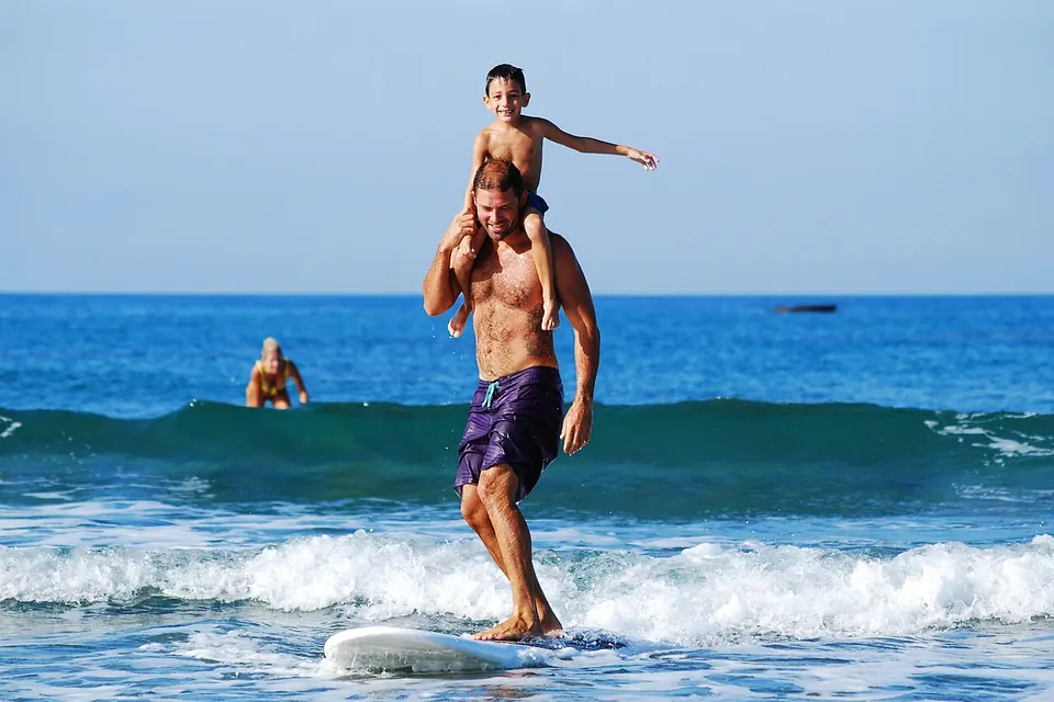 Pai caminhando na água do mar com o filho pequeno nos ombros, de braços abertos