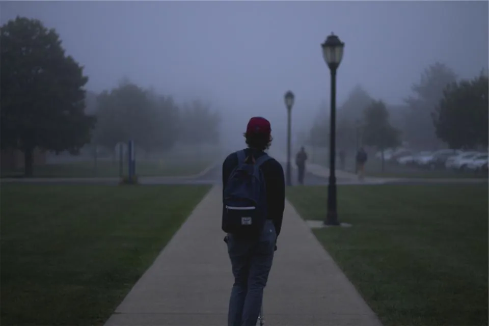 Estudante de mochila caminhando sozinho por uma trilha num parque com neblina