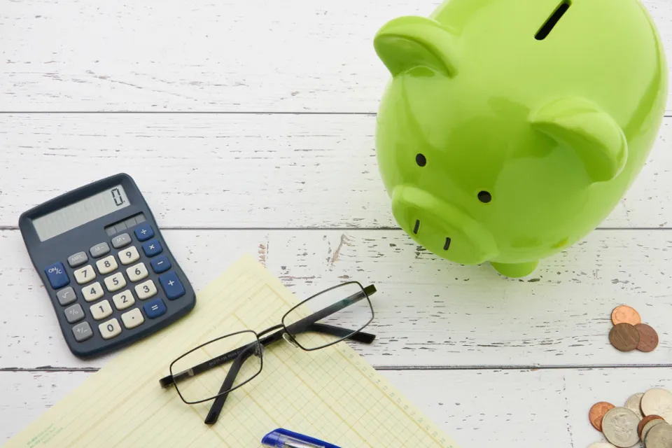 Cofrinho verde em forma de porquinho ao lado de uma calculadora, óculos e um bloco de anotações