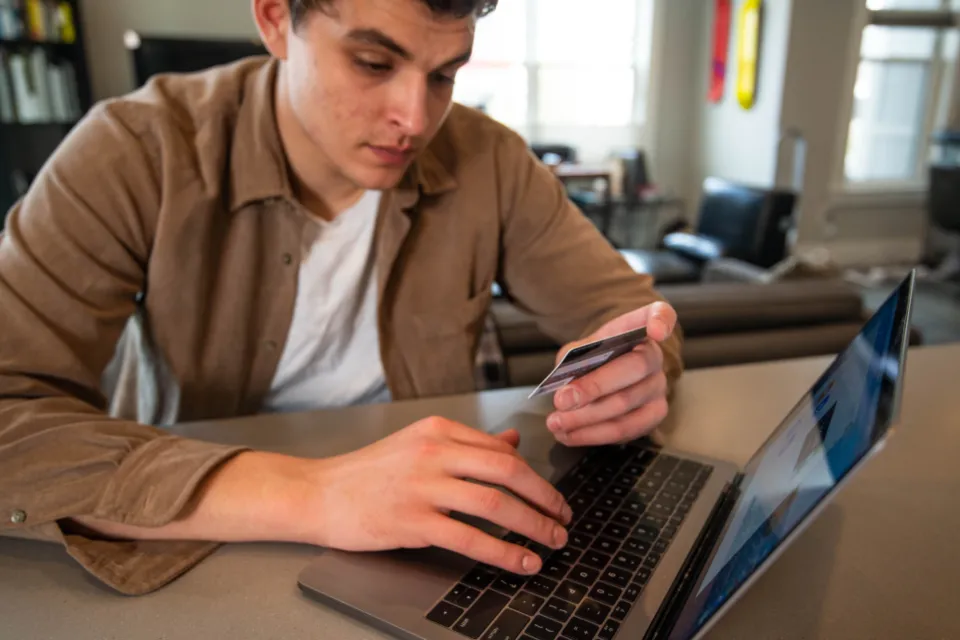 Jovem segurando um cartão de crédito enquanto faz uma compra num notebook
