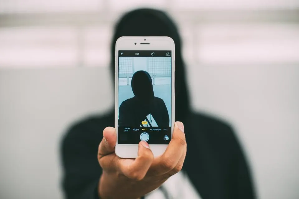 Pessoa de moletom com o rosto escondido atrás de um celular que mostra a própria foto