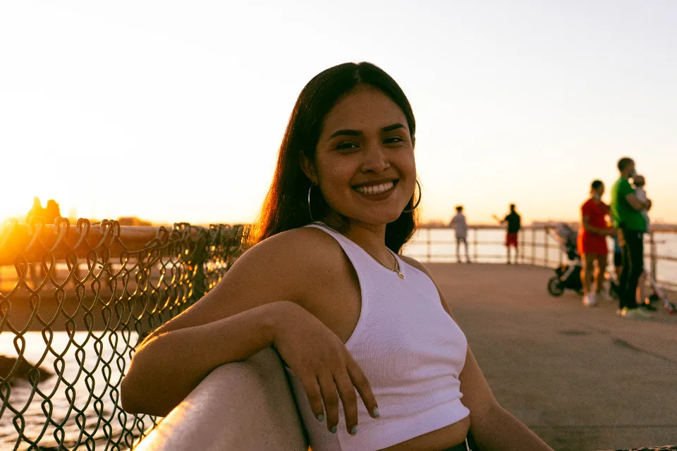Jovem sorrindo sentada à beira de um rio ao pôr do sol