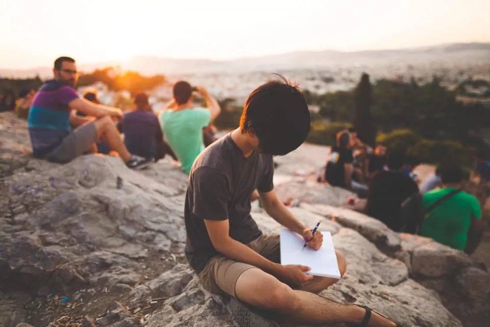 Grupo de jovens sentados em pedras ao ar livre, um deles escrevendo em um caderno