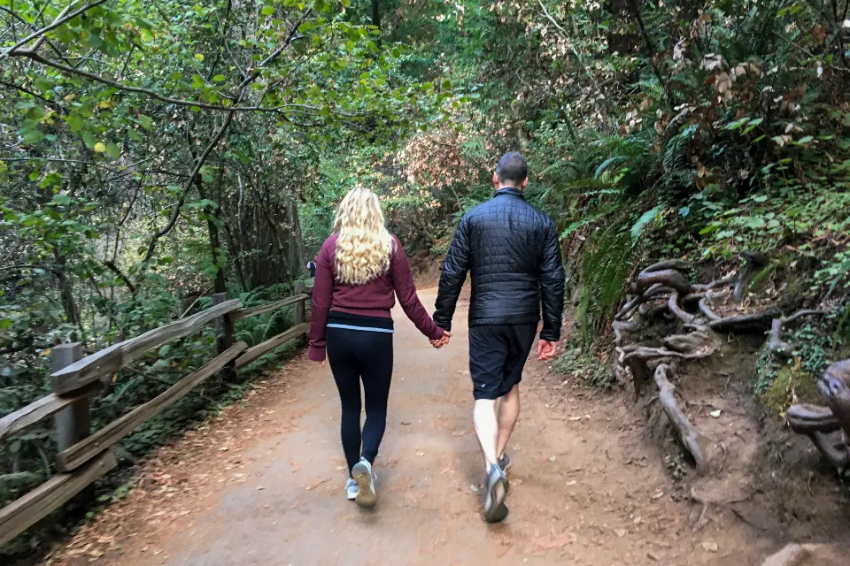 Casal de mãos dadas caminhando de costas por uma trilha no meio da mata