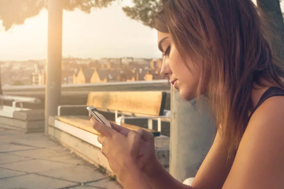 Jovem sentada sozinha olhando para o celular, com a cidade ao fundo