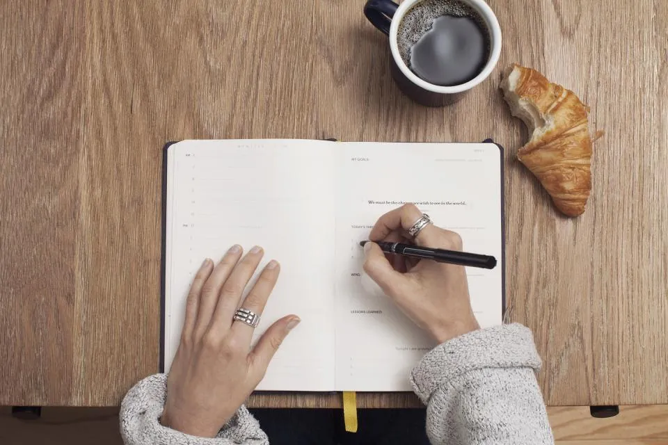 Pessoa escrevendo em um caderno sobre a mesa, ao lado de uma xícara de café